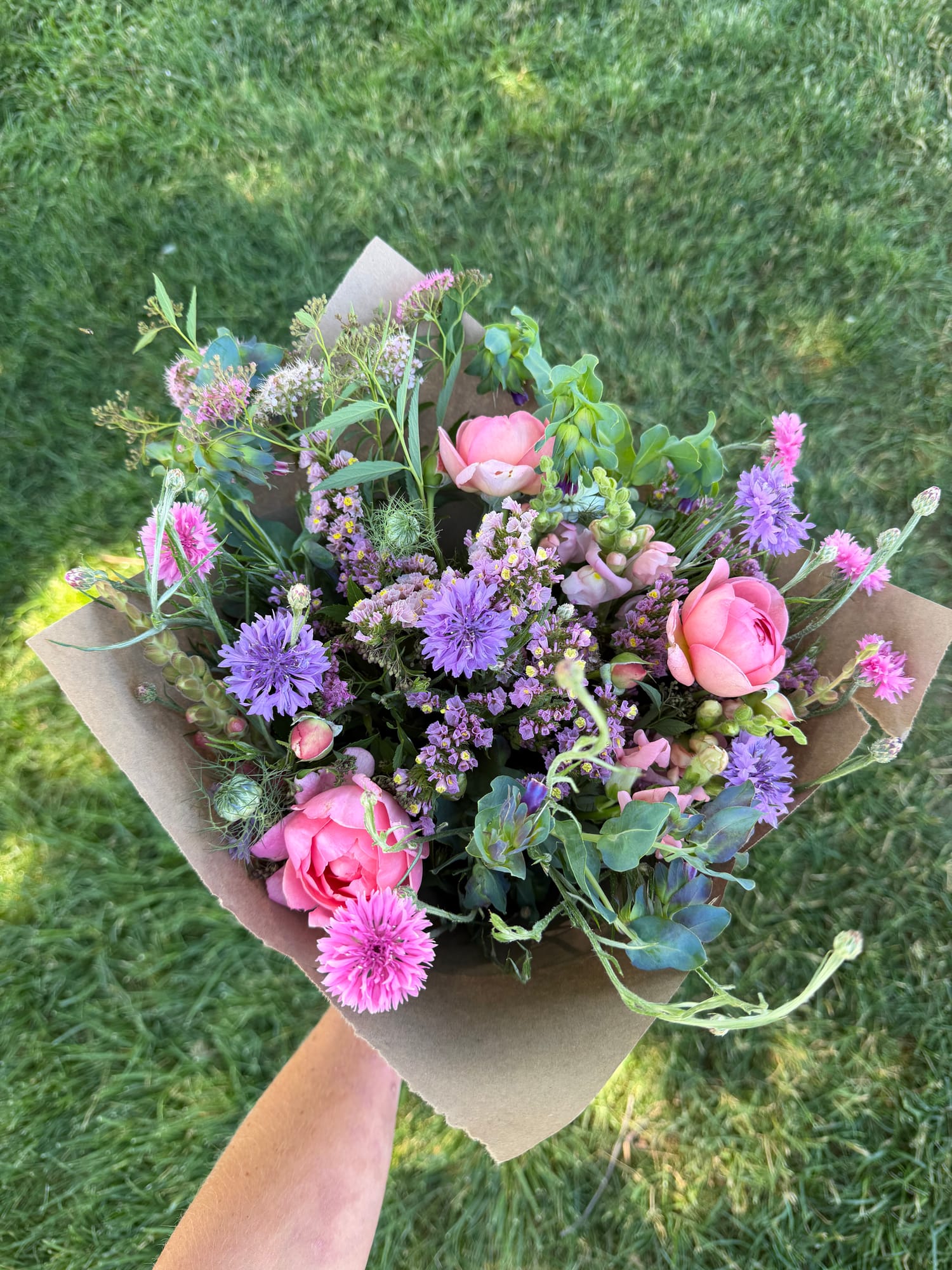 A soft bouquet of pink roses, purple cornflowers, and statice wrapped in kraft paper