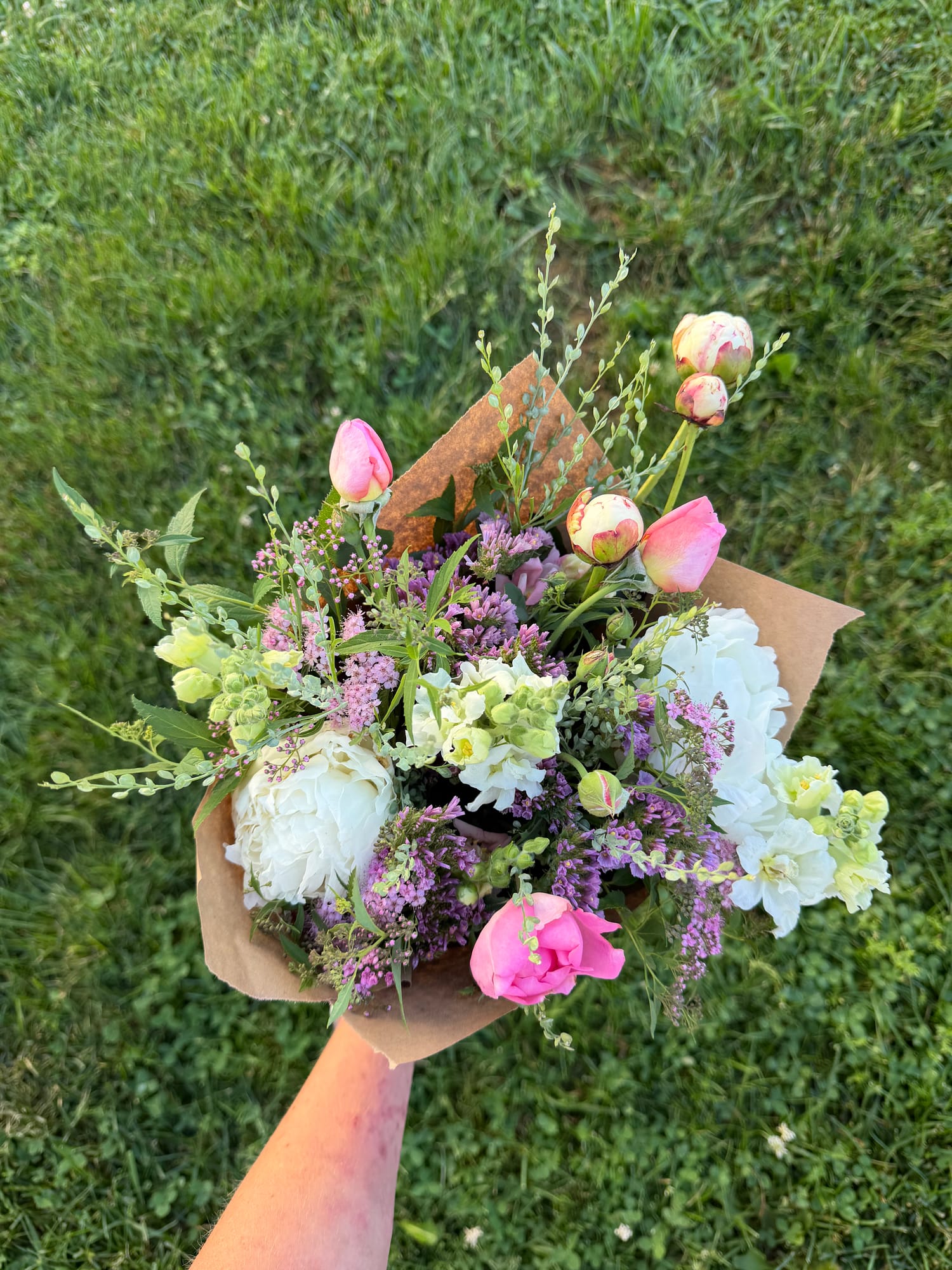 A handmade bouquet of white peonies, pink roses and tulips, and purple statice wrapped in kraft paper
