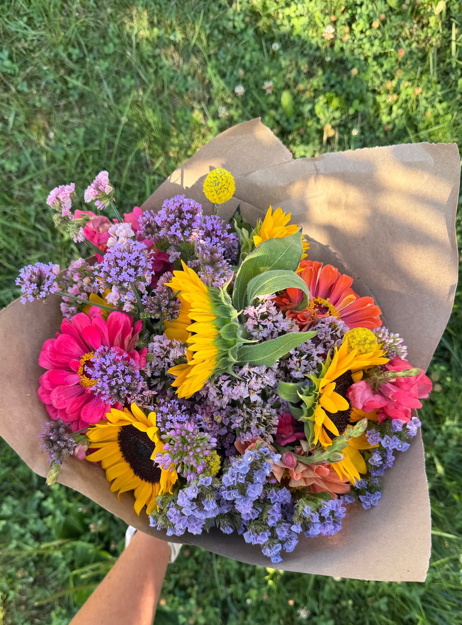A bright bouquet of sunflowers, pink zinnias, and purple verbena wrapped in kraft paper