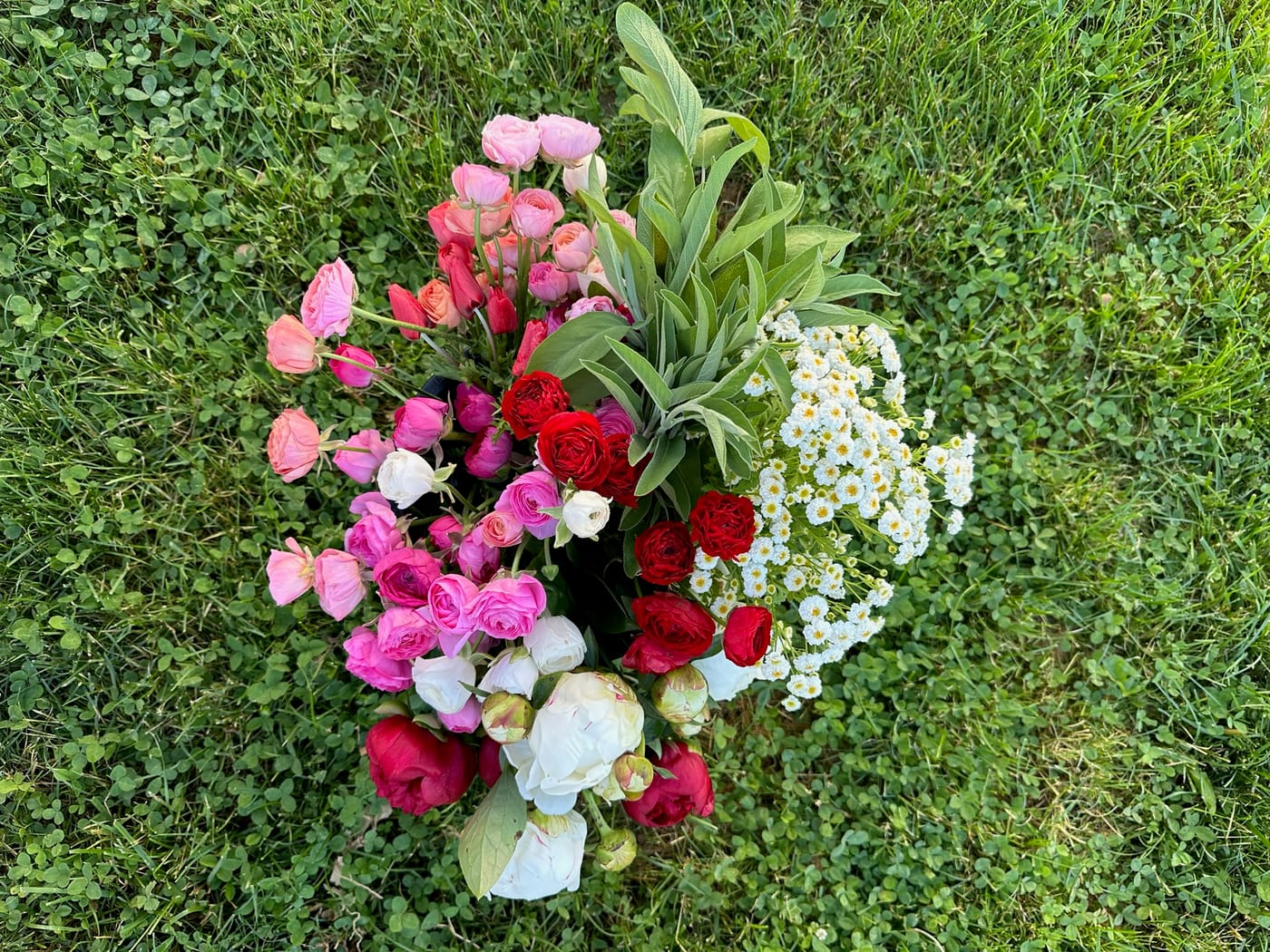 An overhead bouquet of pink and red ranunculus, roses, and peonies on the grass